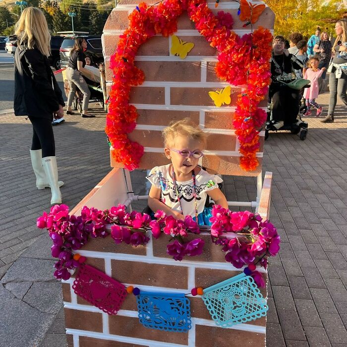Little girl smiling inside a colorful Encanto themed floral booth wheelchair costume for Halloween.