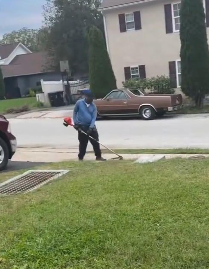 A neighbor using a string trimmer on the edge of a lawn with a car in the background A neighbor using a string trimmer on the edge of a lawn with a car in the background