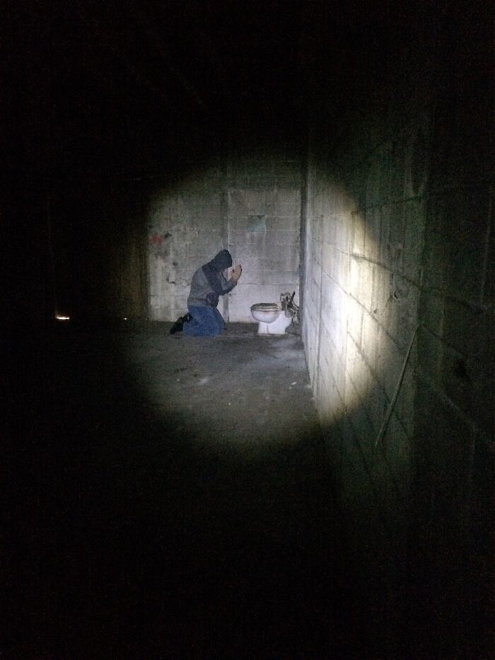 Person kneeling by broken toilet in dark room lit by flashlight beam