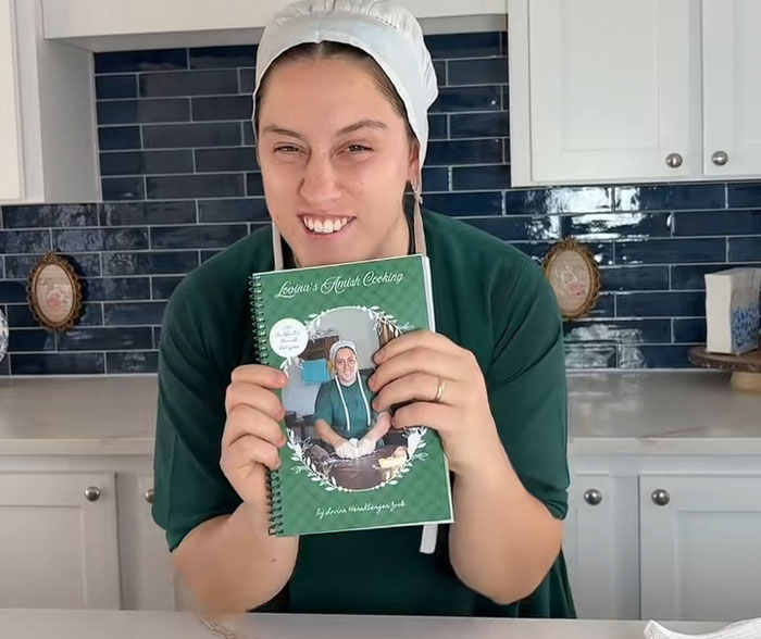 Lovina Zook holding her cookbook titled Lovina's Amish Cooking while smiling in her modern kitchen. Lovina Zook holding her cookbook titled Lovina's Amish Cooking while smiling in her modern kitchen.