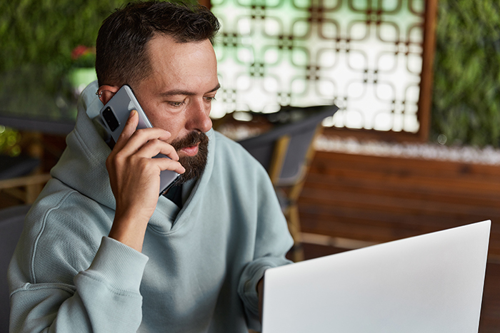 A bearded man wearing a hoodie talking on his smartphone while looking at a laptop. A bearded man wearing a hoodie talking on his smartphone while looking at a laptop.