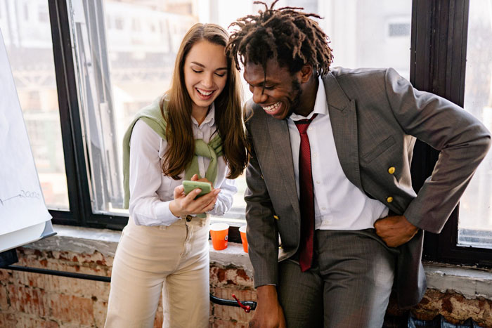 Man and woman smiling while looking at a smartphone in a casual office setting Man and woman smiling while looking at a smartphone in a casual office setting