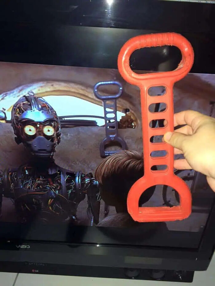 A hand holding a red plastic toy tool next to a screen showing C-3PO and similar movie props