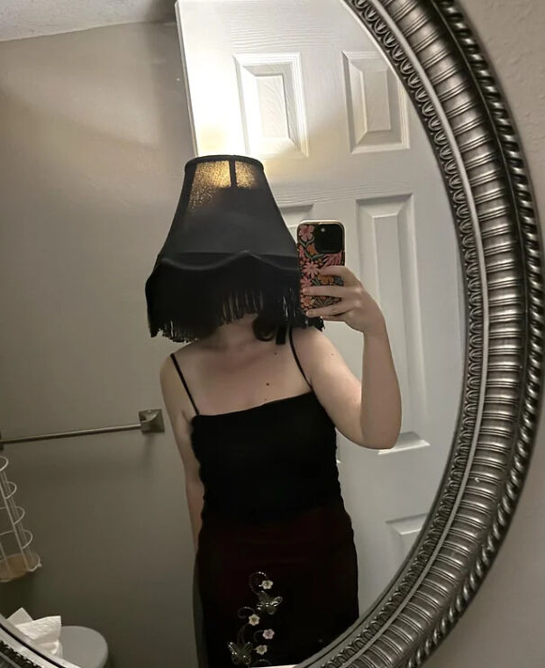 A woman taking a mirror selfie wearing a lampshade on her head for a cool Halloween costume idea.