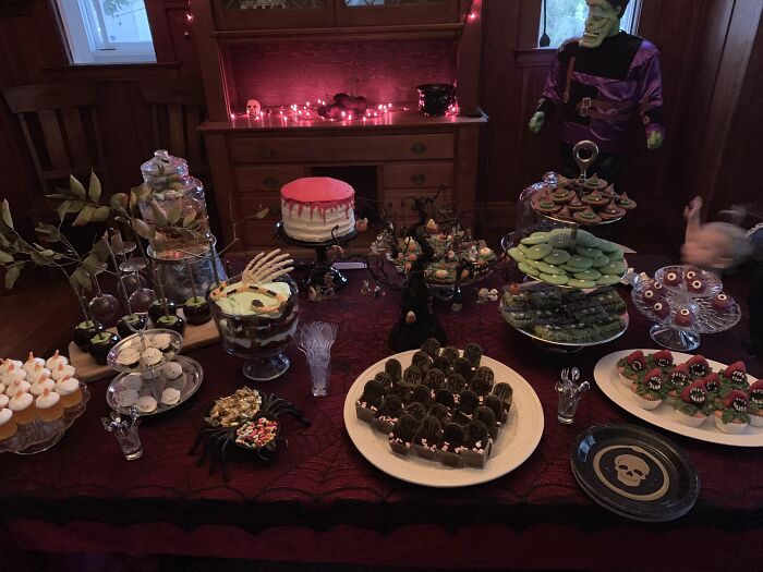 Party table filled with spooky Halloween foods including grave cookies, skull decor, and themed treats.