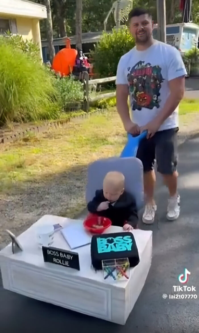 A toddler dressed in a Boss Baby Halloween costume sitting at a mini office desk pushed in a stroller