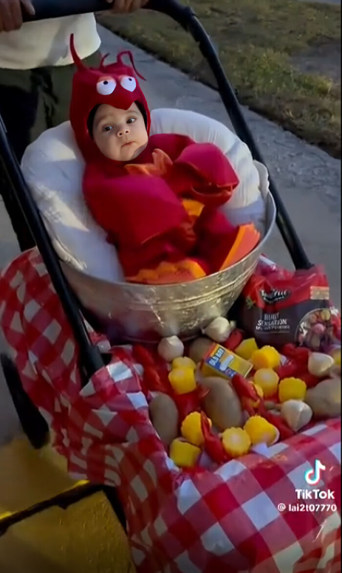 A baby dressed in a red lobster Halloween costume sitting inside a pot filled with corn and potatoes
