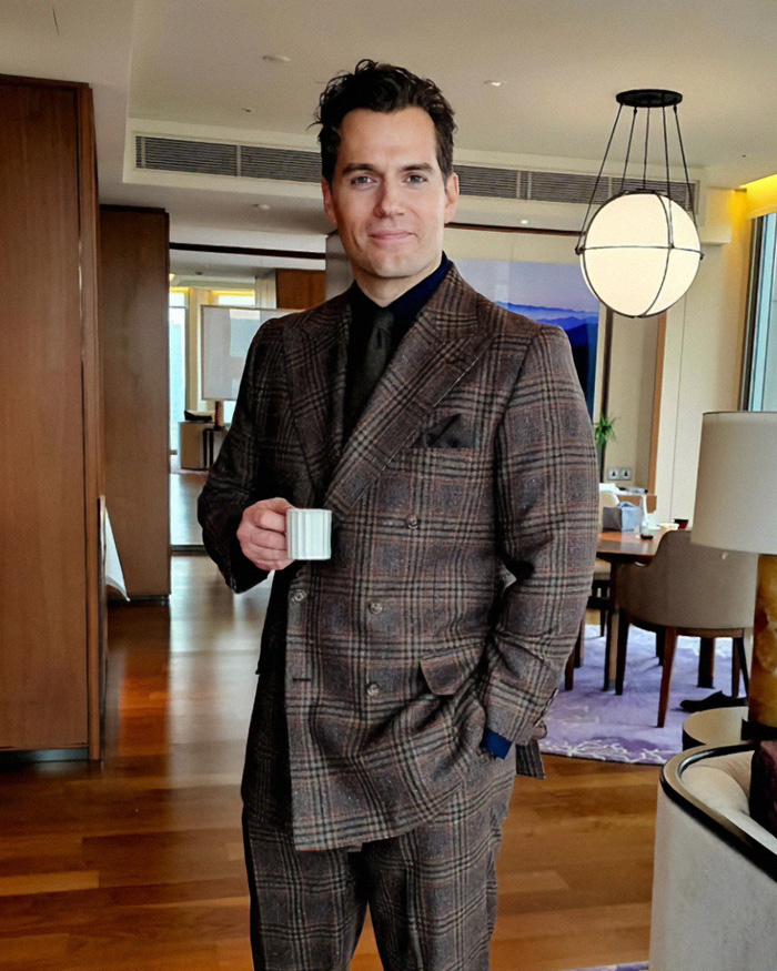 Actor Henry Cavill looking sharp in a brown plaid suit while holding a small cup, highlighting his transformation.