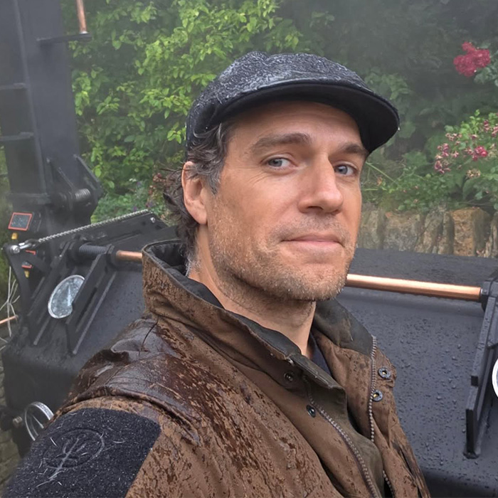 Henry Cavill in a flat cap and brown jacket, representing Henry Cavill's Five-Year Transformation.