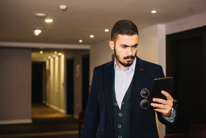 Man in hotel corridor looking puzzled at phone, related to hotel room booking mistake Man in hotel corridor looking puzzled at phone, related to hotel room booking mistake