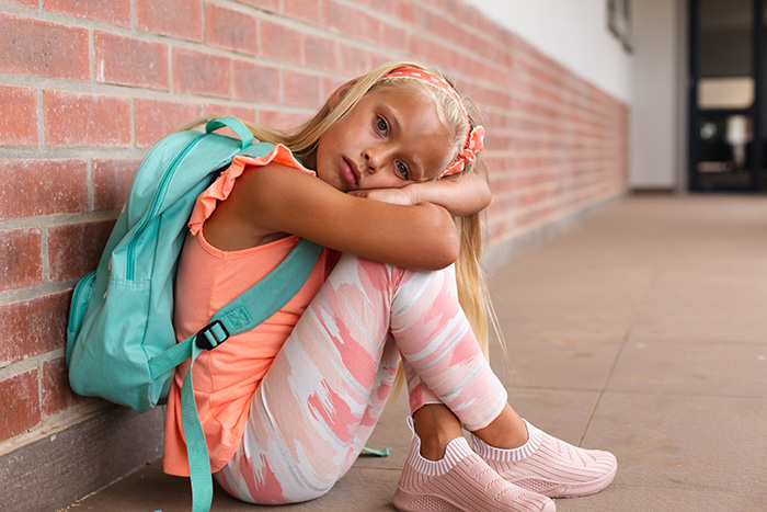 A sad young girl with a backpack sitting alone against a brick wall, feeling left at school. A sad young girl with a backpack sitting alone against a brick wall, feeling left at school.