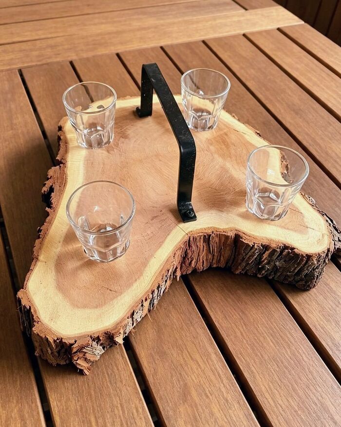 An artistic wooden creations tray made from a tree slice holding four clear shot glasses with a metal handle.