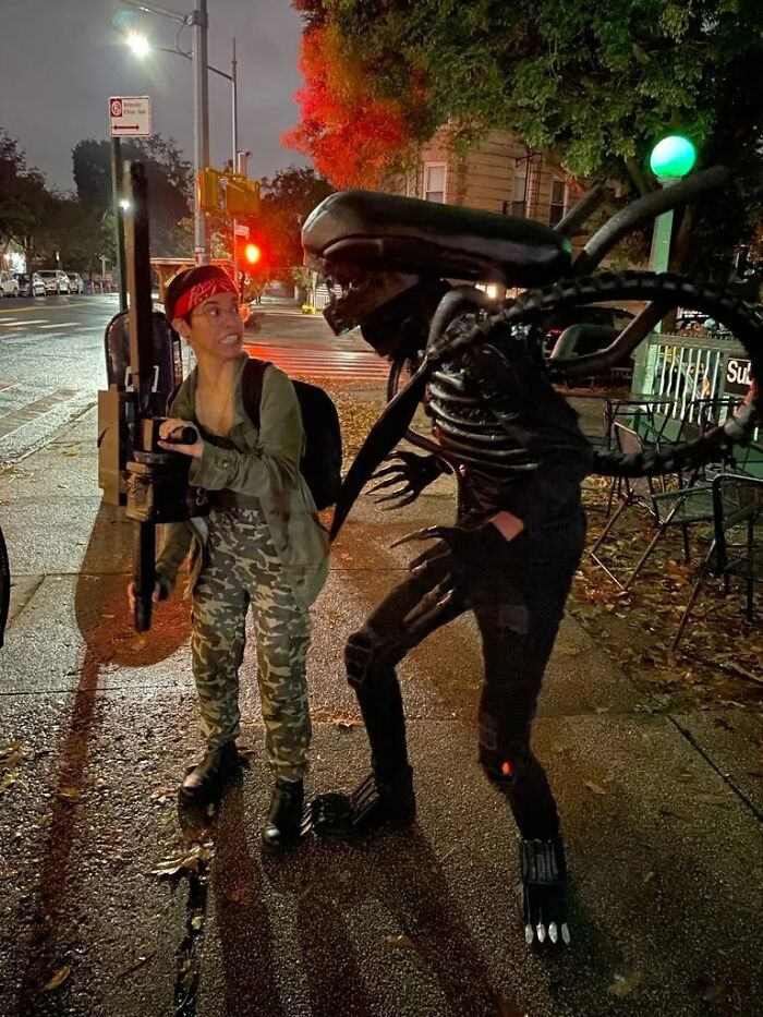 Funny Halloween couples costume featuring Ripley and a Xenomorph alien standing on a city sidewalk at night