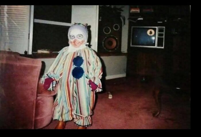 A child standing in a dimly lit living room wearing a weird and creepy clown costume.