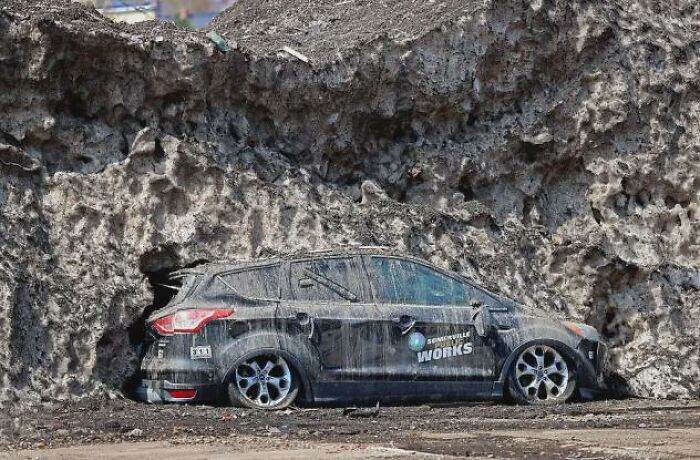 A dirty car parked under a massive wall of melted snow in weird photos.