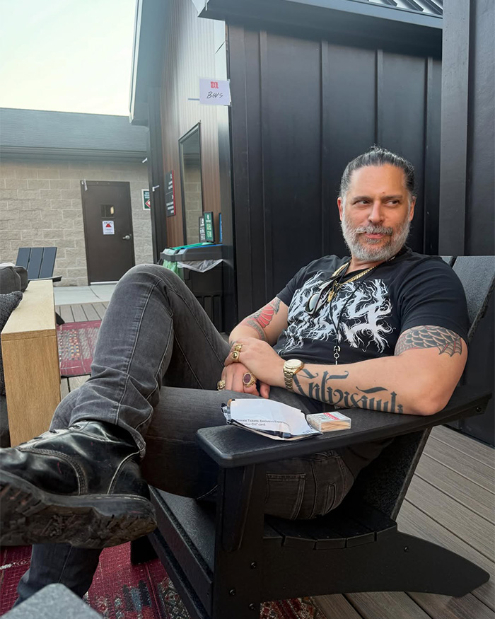 Joe Manganiello sitting on an outdoor chair, wearing a black graphic t-shirt and grey jeans.