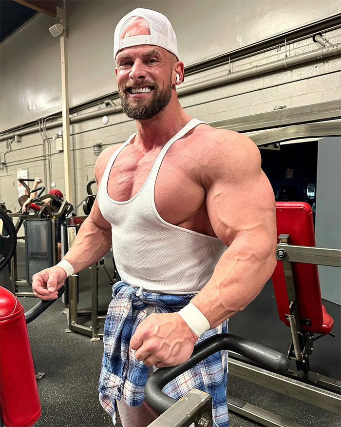 Joey Swoll smiling in a white tank top showing his muscular physique at the gym Joey Swoll smiling in a white tank top showing his muscular physique at the gym