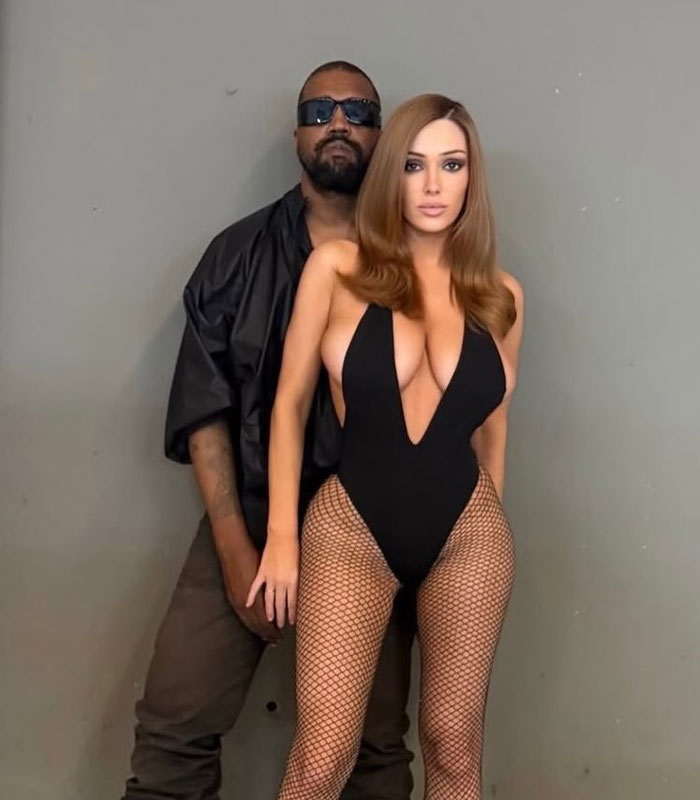Kanye West standing behind Bianca Censori as she poses in a revealing black bodysuit and fishnet tights.