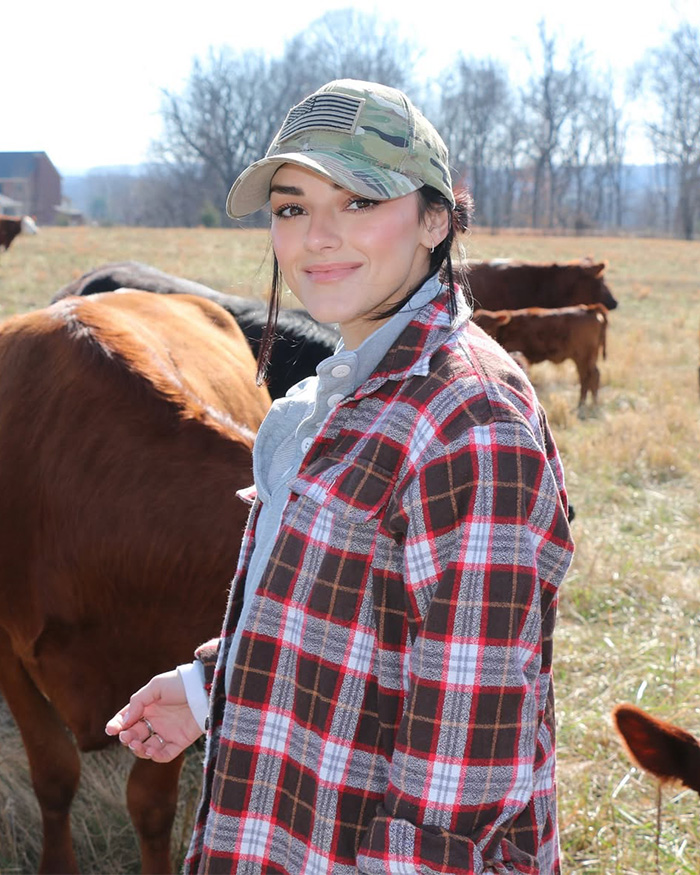 Kash Patel fiance&eacute; wearing plaid shirt and camo hat standing near cows in a field