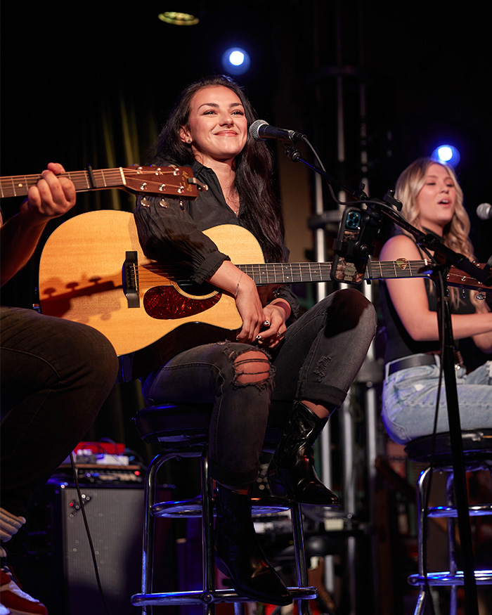 Kash Patel's fiance&eacute; smiling while playing an acoustic guitar on stage under warm lighting.