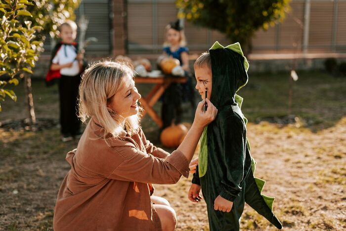 Woman applying face paint to a child wearing a green dinosaur costume for Halloween
