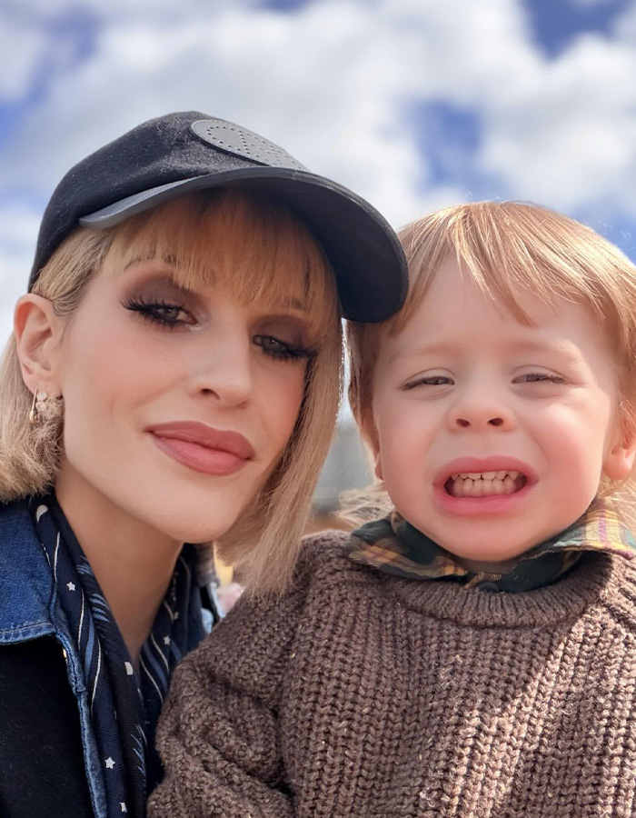 Kelly Osbourne posing in a black cap alongside her young child wearing a cozy brown sweater.