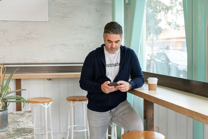 A middle-aged man standing near a window looking at his phone, representing adult children of narcissistic parents