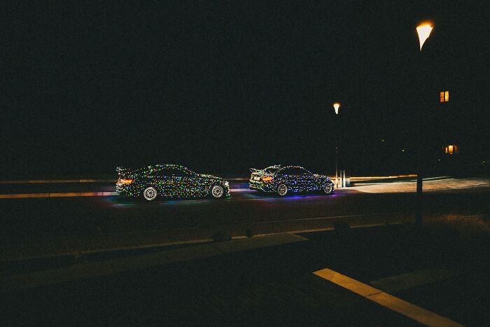Two sports cars illuminated with colorful Christmas lights at night, creating a Hollywood-style scene.