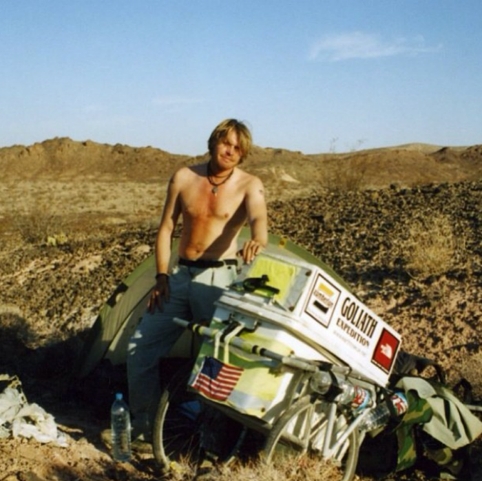 A man goes for a walk in 1998, posing shirtless next to his Goliath Expedition cart in a desert setting. A man goes for a walk in 1998, posing shirtless next to his Goliath Expedition cart in a desert setting.