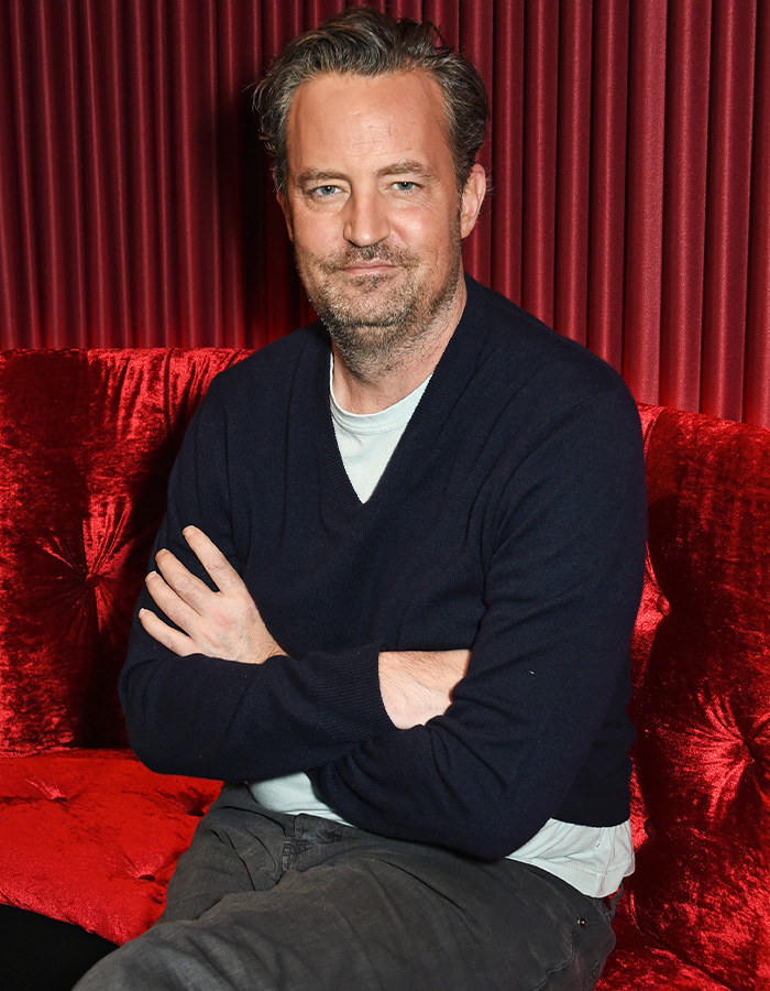 Actor Matthew Perry wearing a dark sweater seated on a red velvet couch.