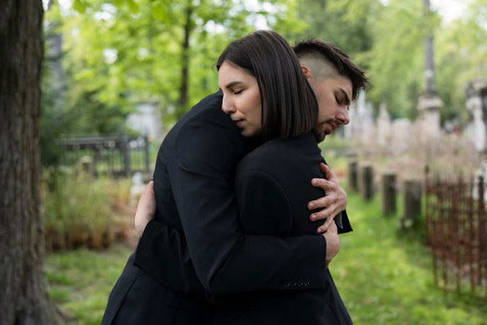 An image of a young man and woman hugging in grief at a cemetery, illustrating a teen works to buy first car backstory. An image of a young man and woman hugging in grief at a cemetery, illustrating a teen works to buy first car backstory.