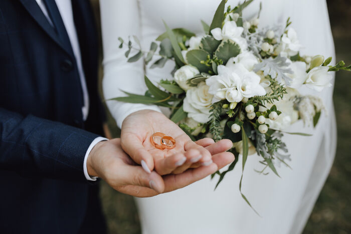 A bride and groom holding wedding rings and a floral bouquet