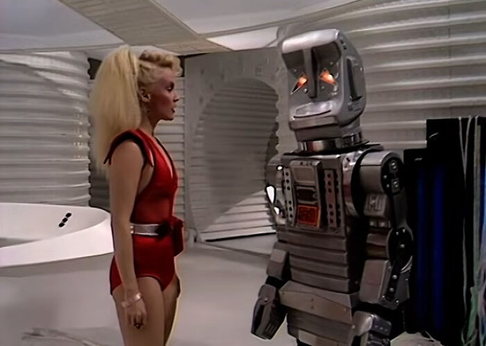 A woman in a red outfit talking to a vintage robot, symbolizing intelligent insults from sci-fi