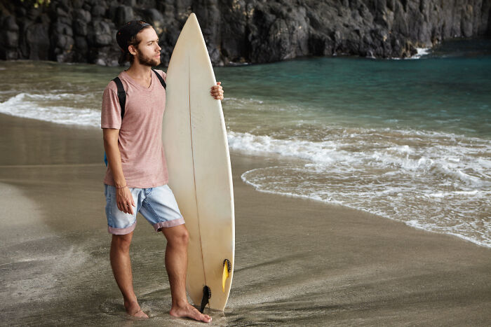 A man with a surfboard standing on the beach, unaware of potential intelligent insults.