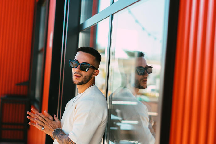 A man in sunglasses leaning against a window, delivering intelligent insults with a cool expression