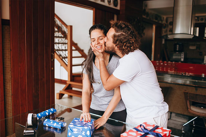 A man lovingly kissing a woman on the cheek while she opens a gift, illustrating a guy pursuing ex story.