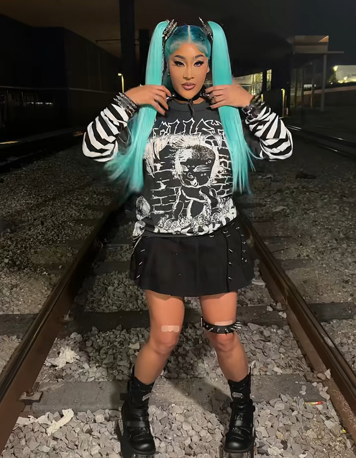 Natalie Nunn wearing bright blue pigtails and a black gothic outfit outdoors on train tracks.