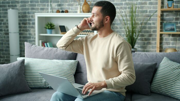 A man talking on the phone while using a laptop on his couch, stirring up drama in tiny communities.