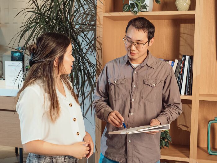 A man with glasses holding a clipboard talking to a woman in a tiny community office.