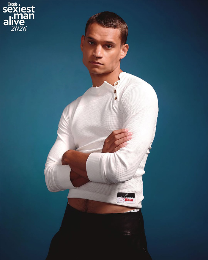 Man posing in a white long-sleeved shirt during a Sexiest Man Alive photoshoot against a blue background