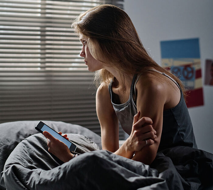A sad teen girl sitting on her bed holding a phone in a dimly lit room