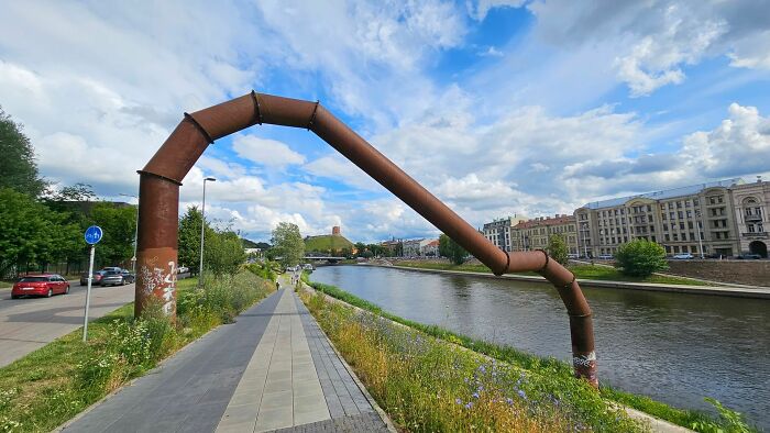 Large rusted metal pipe arching over a riverside pedestrian path as a public art piece.