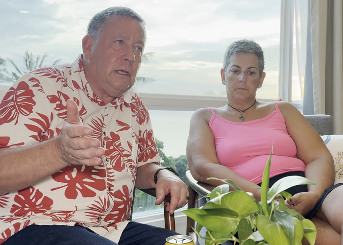 A man in a floral shirt speaking animatedly alongside a woman indoors, discussing permanent cruise life.