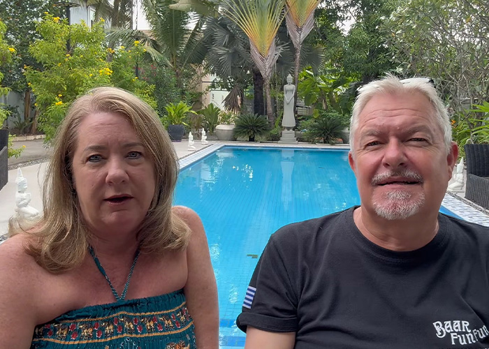 A middle-aged couple seated outdoors near a swimming pool, speaking about their permanent cruise experience.
