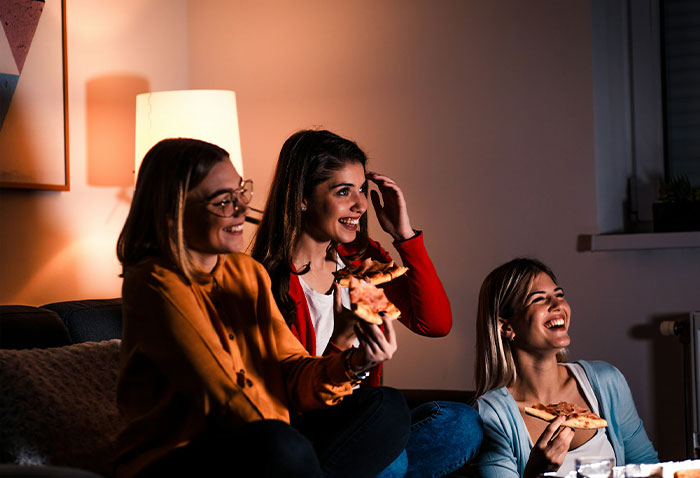 Three roommates eating pizza while watching TV in a cozy living room. Three roommates eating pizza while watching TV in a cozy living room.