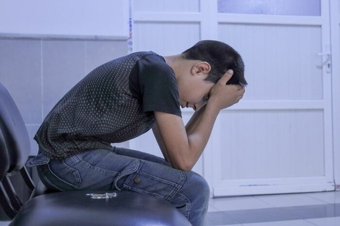 Stressed boy sitting alone in waiting room with head in hands, showing embarrassment