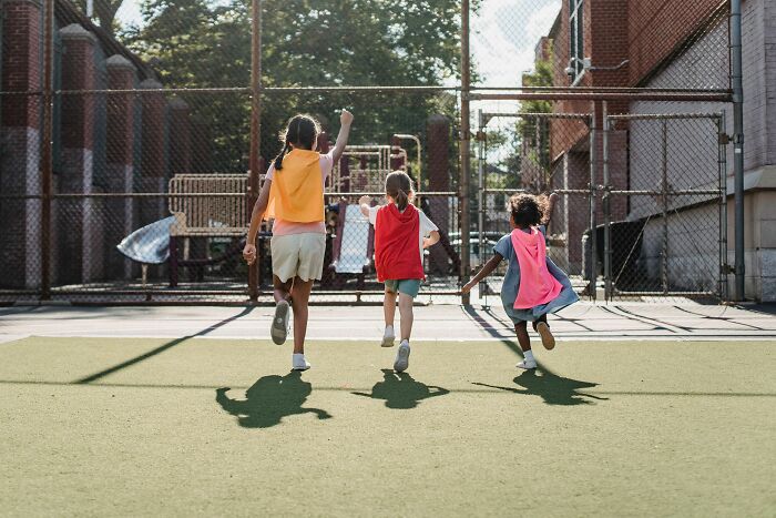 Three children wearing capes running together in a tiny community school playground.