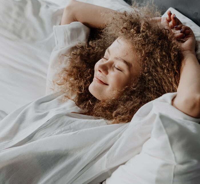 Happy young woman with curly hair relaxing in bed, expressing self-care tips women wish they knew sooner.