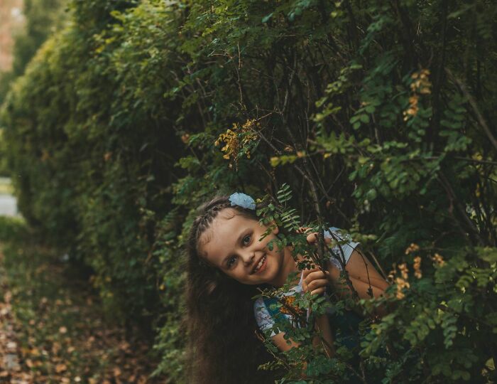 A young girl peeking through green bushes in a funny and playful moment