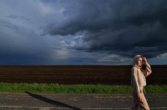 A girl in a hoodie walking by a field under dark storm clouds, sensing an urgent feeling to leave